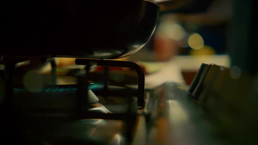 A hand ignites a gas burner on a stainless steel stove with a blue flame appearing under a black pan