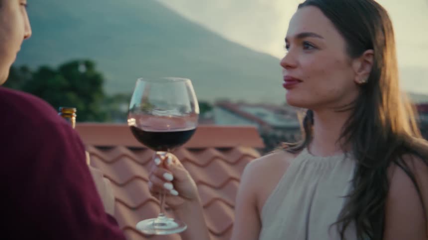 A beautiful woman swirls her red wine and enjoys a romantic conversation with her partner on a scenic rooftop during a warm sunset