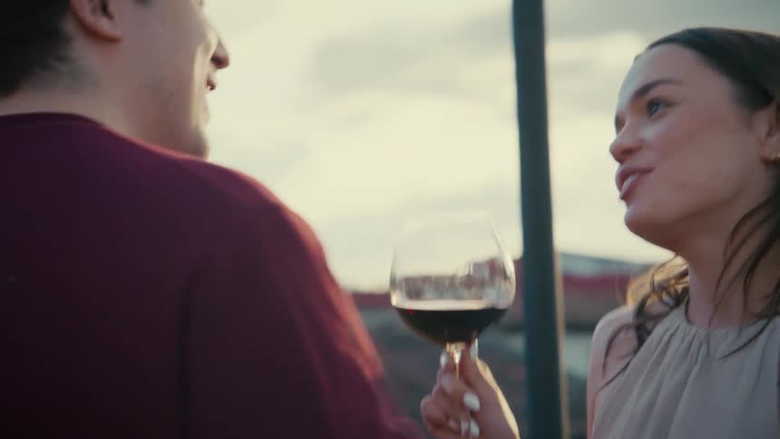 A lovely young woman smiles and chats with her date, holding a glass of red wine on a scenic rooftop during a warm sunset