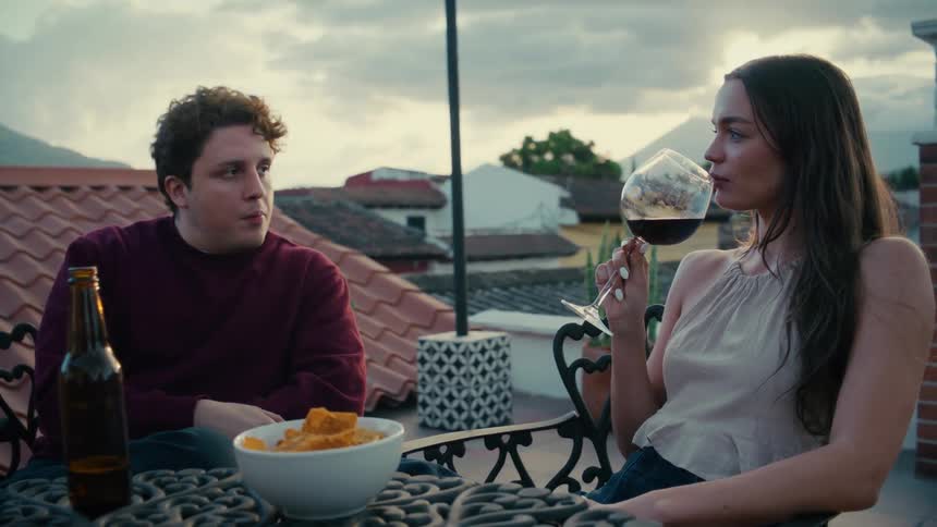 A relaxed young couple shares drinks, snacks, and conversation on a beautiful rooftop terrace with mountains in the background