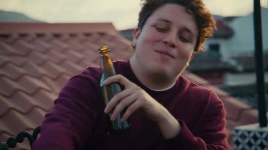 A man and woman enjoy drinks and conversation on a rooftop patio overlooking a scenic town during a beautiful sunset