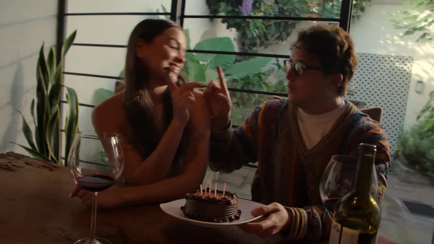 A young couple celebrates a birthday by lighting candles on a chocolate cake, blowing them out, and playfully eating it together with red wine