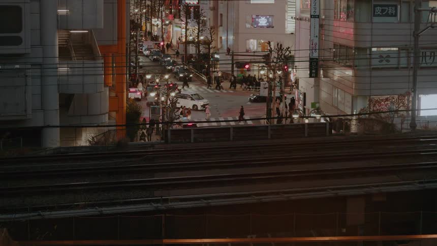 A high-angle shot of trains passing by with a busy Tokyo street illuminated by city lights in the background at night