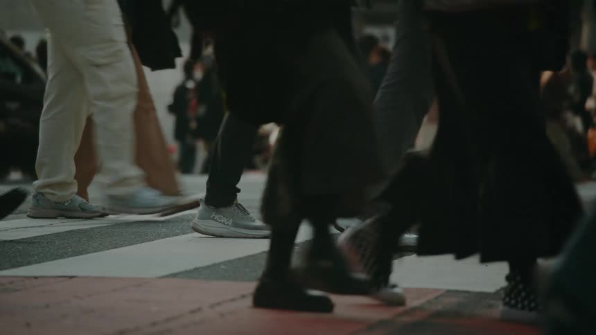 A low-angle slow-motion shot captures the feet of a diverse crowd of anonymous pedestrians as they cross a busy city street