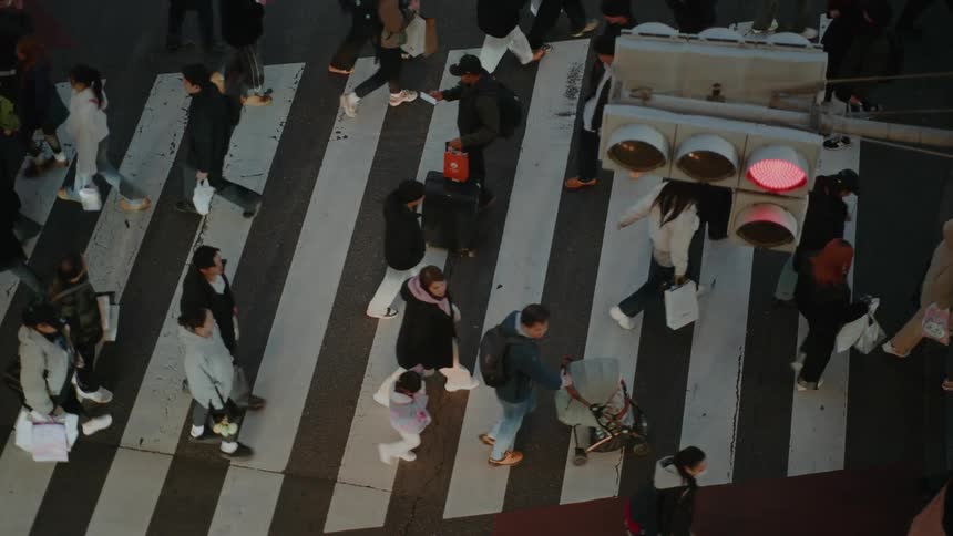 A slow-motion, high-angle shot reveals a crowd of pedestrians crossing a busy street in a modern city at dusk
