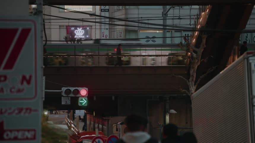 A long shot captures the layered movement of a busy Tokyo street at night, with pedestrians on an overpass and a train speeding by