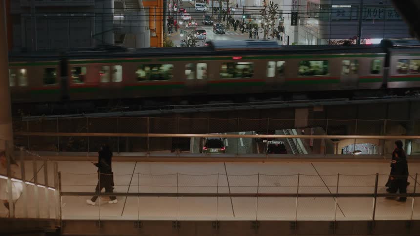 A high-angle view of pedestrians on an illuminated walkway as a train passes by, with busy city traffic visible below at night