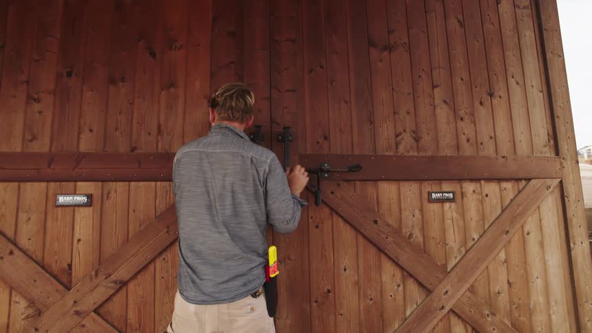 A man with a tool on his hip walks up to a large wooden barn, unlatches the doors, and enters in slow motion