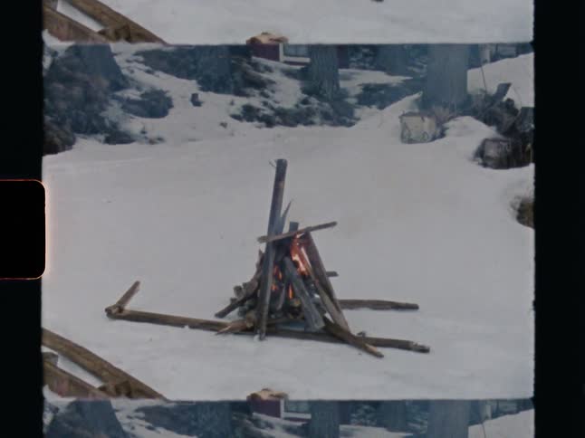 A rustic campfire burns brightly in a teepee formation surrounded by snow in this vintage film clip