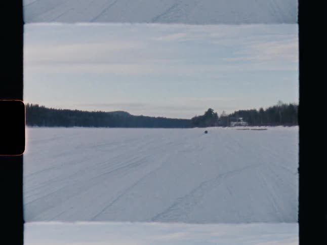 A snowmobile drives away across a vast, frozen lake in a winter landscape, captured on vintage film