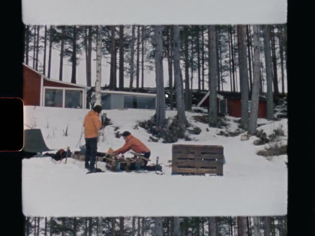 Vintage 8mm film shows two men in winter jackets preparing equipment in a snowy forest landscape