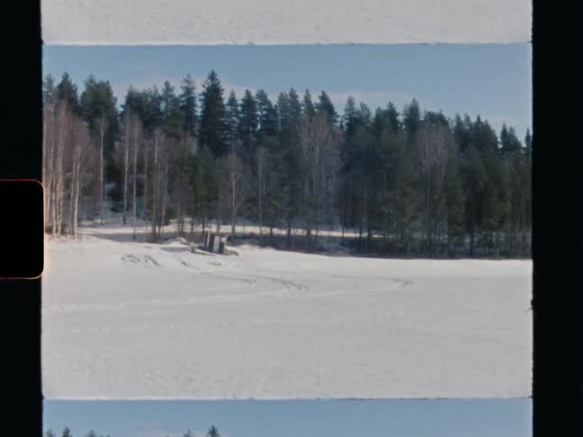A wide, static shot from vintage 8mm film shows a vast, snowy landscape in front of a dense pine forest on a sunny day