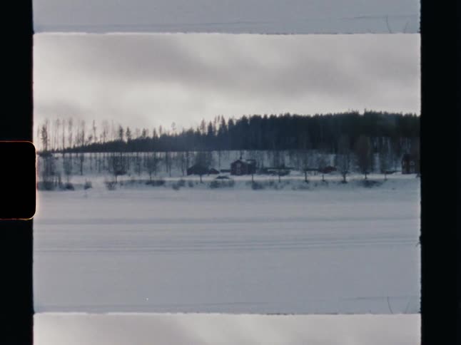 Vintage film captures a still winter landscape with a red cabin across a vast, snow-covered frozen lake