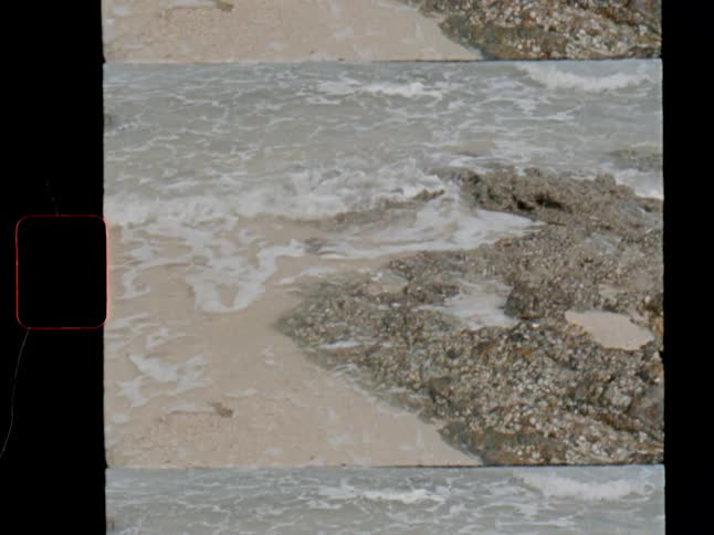 A vintage, slow-motion shot captures gentle ocean waves lapping against a rocky, sandy shoreline