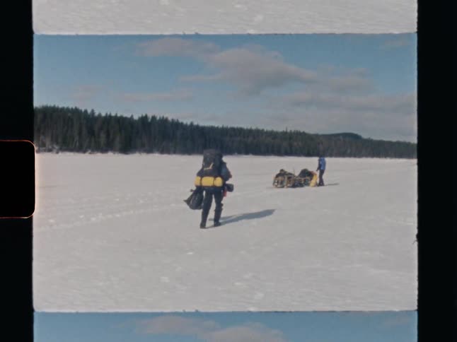 A vintage Super 8 film captures two explorers with backpacks and a sled hiking across a vast, snow-covered frozen lake