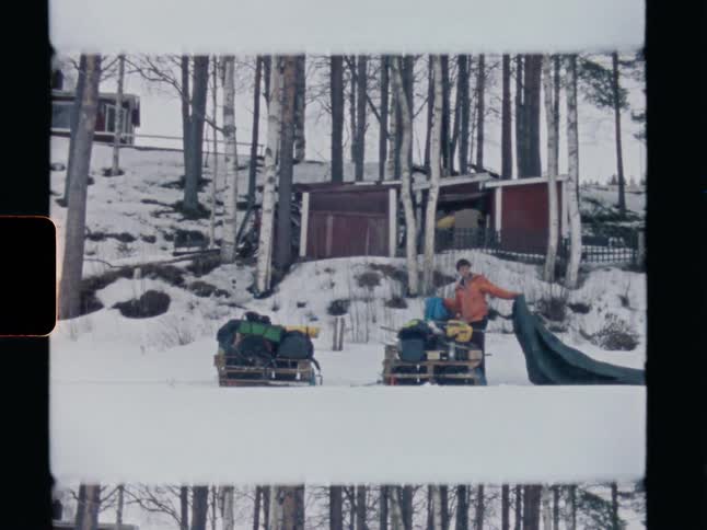 Vintage footage shows a man covering his camping gear on sleds with a tarp in a snowy landscape
