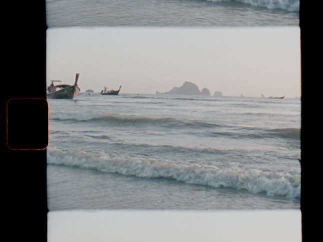 Vintage 8mm film captures the tranquil scene of traditional long-tail boats on the ocean as gentle waves lap the shore