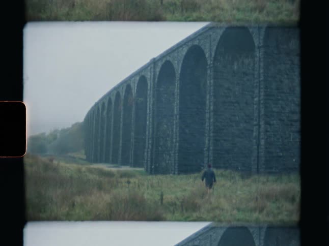 A man walks through a foggy moor towards a towering stone viaduct in this vintage-style footage
