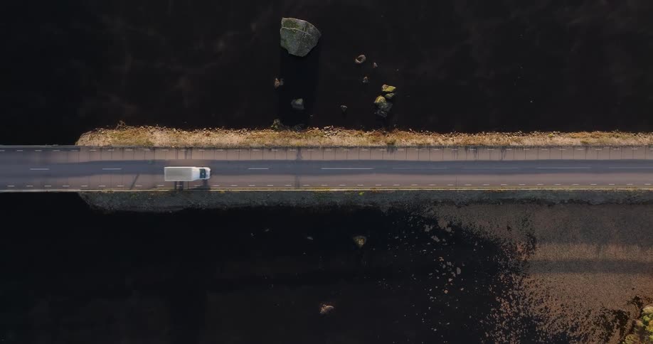 An overhead aerial view captures a white van driving across a scenic causeway surrounded by dark, still water