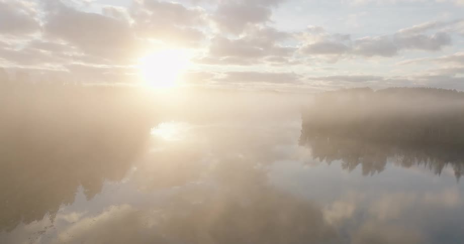 Golden sunrise shines through thick fog over a calm lake, creating a serene and ethereal landscape with reflections of clouds on the water