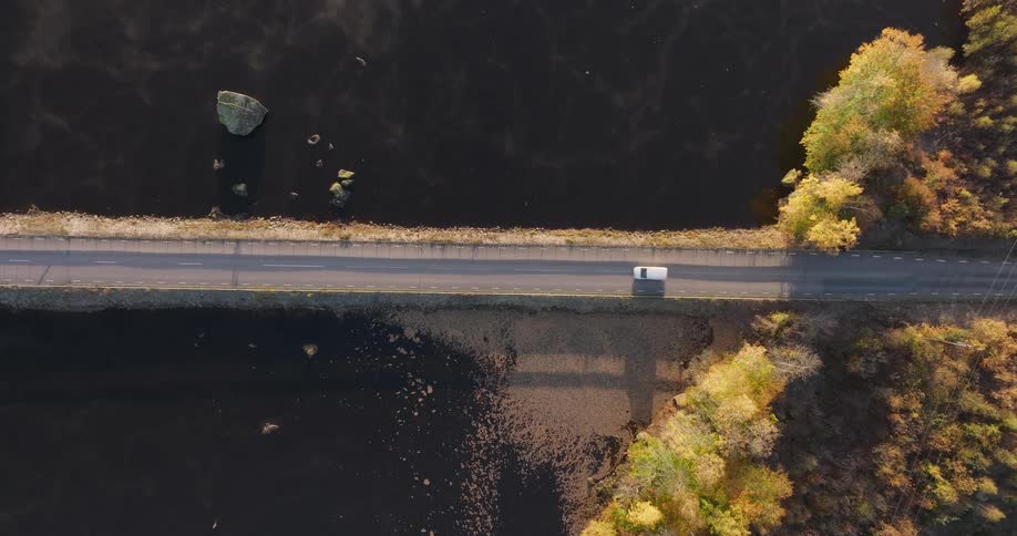 Static top-down aerial view of a white van driving across a causeway over a misty lake surrounded by autumn trees