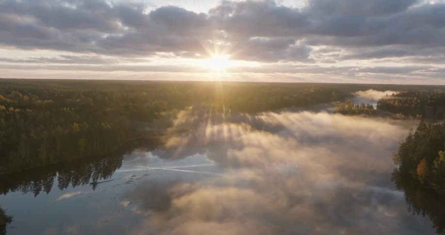 Majestic aerial view of a misty forest lake at sunrise, with beautiful sun rays piercing through the clouds and reflecting on the calm water