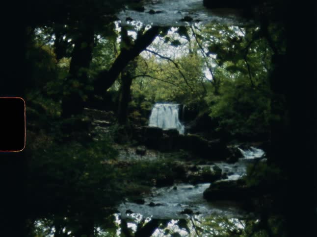 A picturesque waterfall and flowing river are seen through the trees in a lush green forest on grainy, mirrored 8mm film