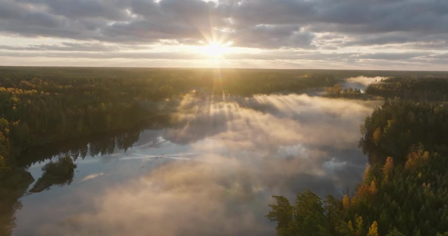 A glorious sunrise casts long rays of light through clouds and mist over a tranquil forest lake in this serene aerial view