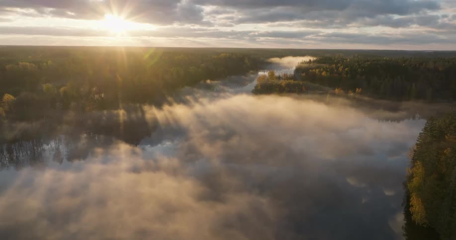 Ethereal aerial footage shows a glorious sunrise casting golden light rays through low-hanging fog over a tranquil river and autumn forest