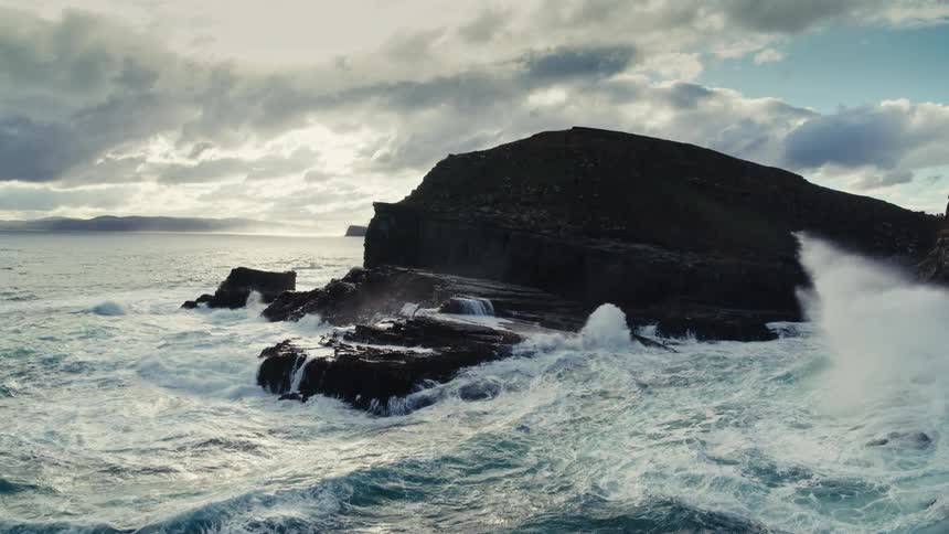 Powerful ocean waves crash in slow motion against a rugged, rocky coastline under dramatic, cloudy skies