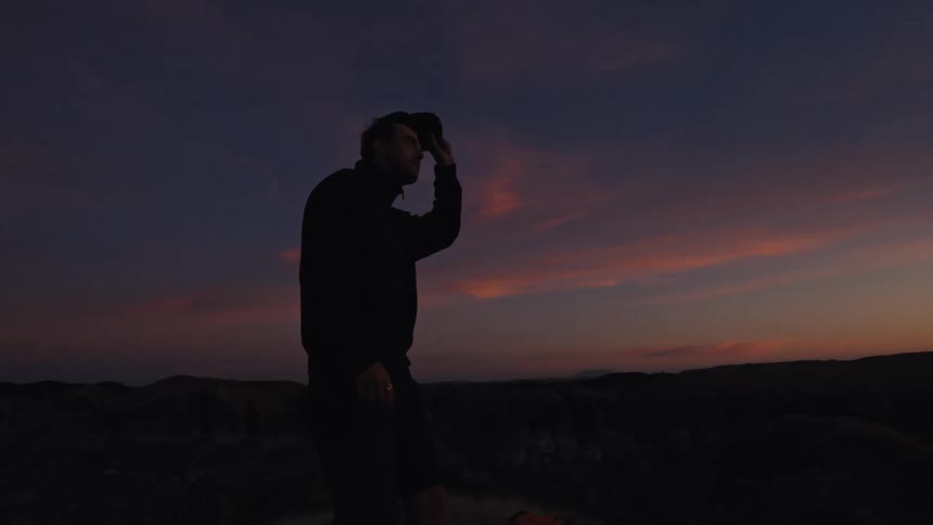 A pensive man silhouetted against a colorful sky takes off his hat to admire the stunning sunset from a rural hilltop