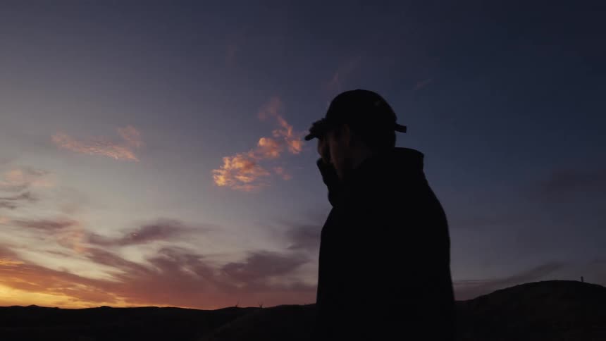 A silhouetted man takes off his cap to watch the colorful sunset over the rolling hills