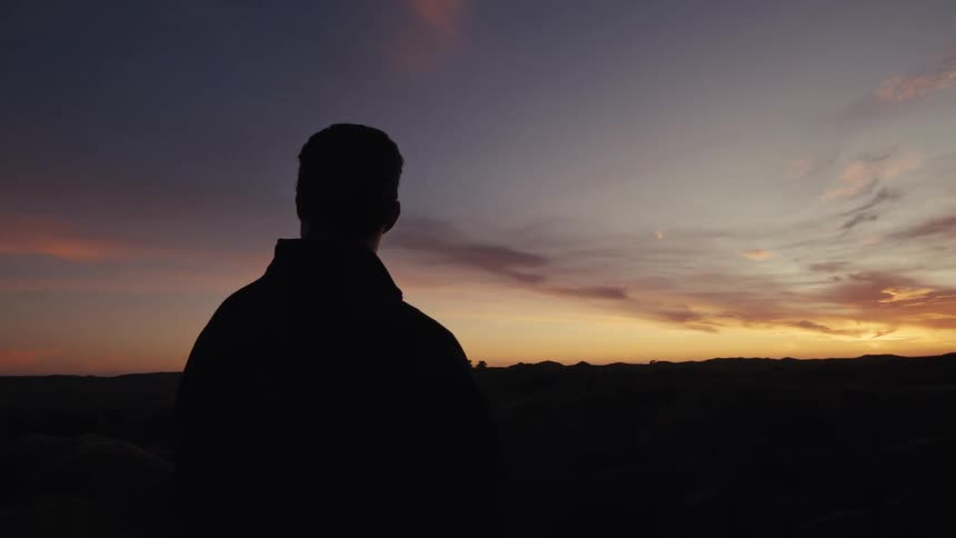 A silhouetted man takes off his hat while watching a beautiful, colorful sunset over a vast, hilly landscape