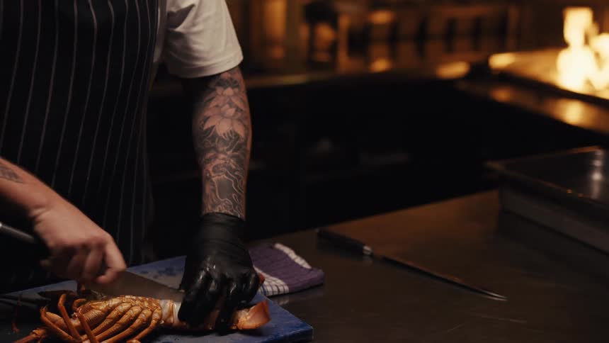 A tattooed chef in black gloves and an apron expertly cuts open a fresh lobster in a professional kitchen with a fire grill in the background