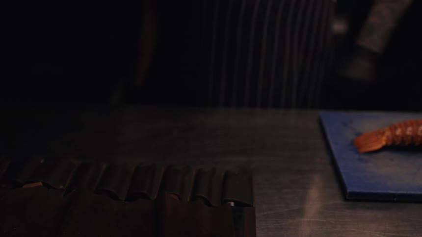 Tattooed chef with black gloves unrolls his professional knife set and begins to prepare a fresh lobster in a dark restaurant kitchen