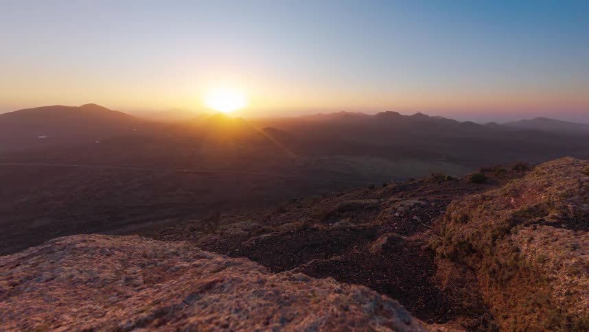 A stunning timelapse captures the golden sun setting behind a vast and rocky volcanic mountain range, casting long shadows across the valley
