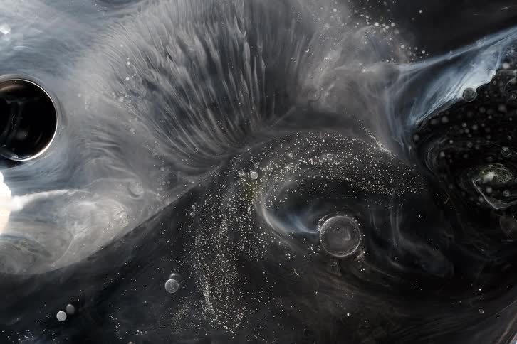 A dark cosmic nebula of black and white paint swirls with shimmering glitter and air bubbles