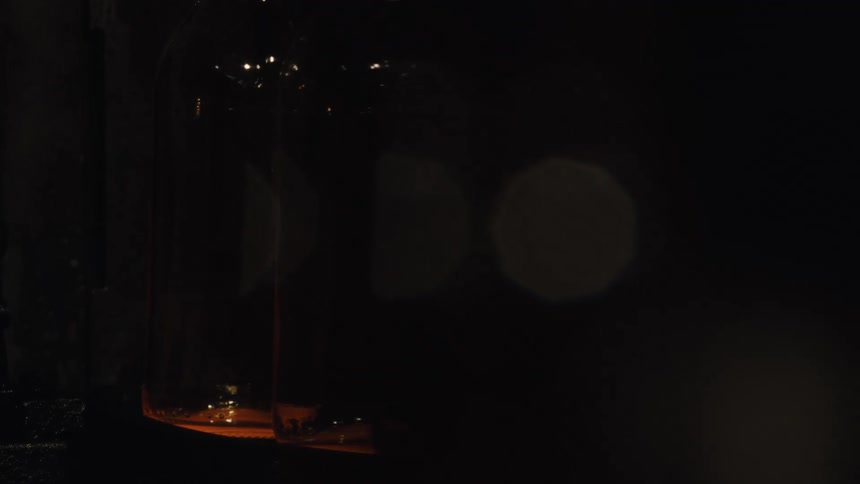 An artistic close-up reveals glowing hot glass bottles moving along a conveyor belt after being molded in a dark factory