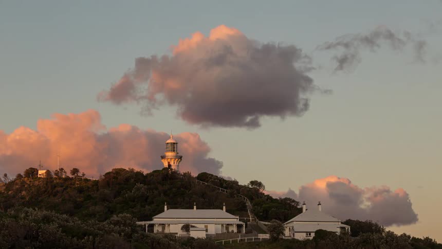 A tranquil time-lapse shows sun-kissed clouds drifting over a classic white lighthouse and keepers' cottages at sunset