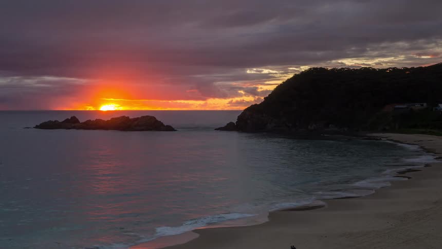 A stunning timelapse shows a vibrant sunrise breaking through dark clouds over a tranquil beach and ocean cove