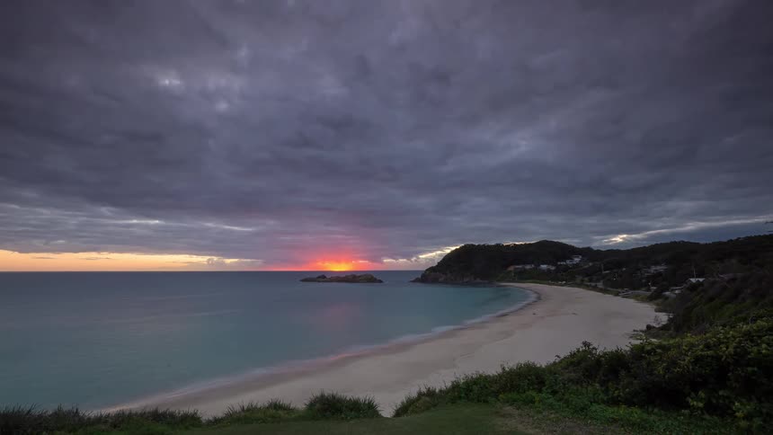 A dramatic time-lapse captures a fiery red sunrise breaking through heavy storm clouds over a tranquil beach cove