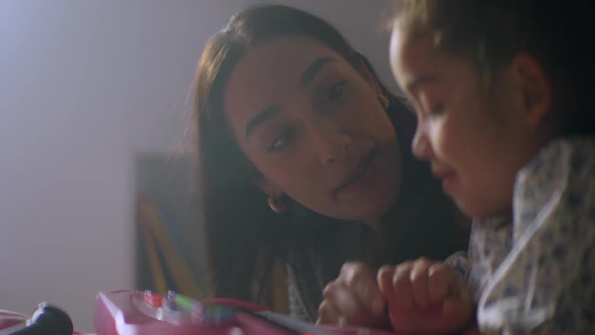 A loving mother proudly watches her young daughter play a pink toy piano in a warmly lit room