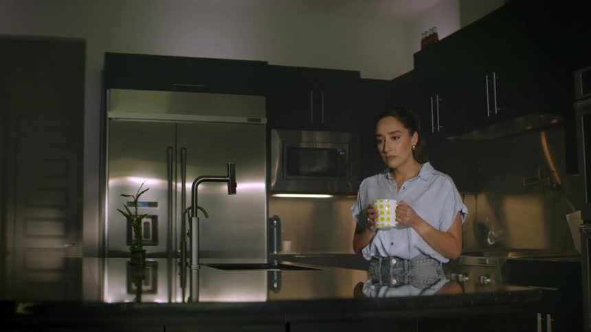 A sad and lonely woman sits on the kitchen counter at night, holding a cup of tea and feeling contemplative