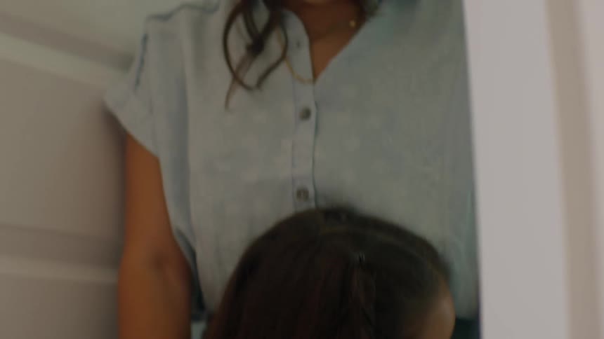 A mother looks down with a somber expression while holding her young daughter in a comforting embrace