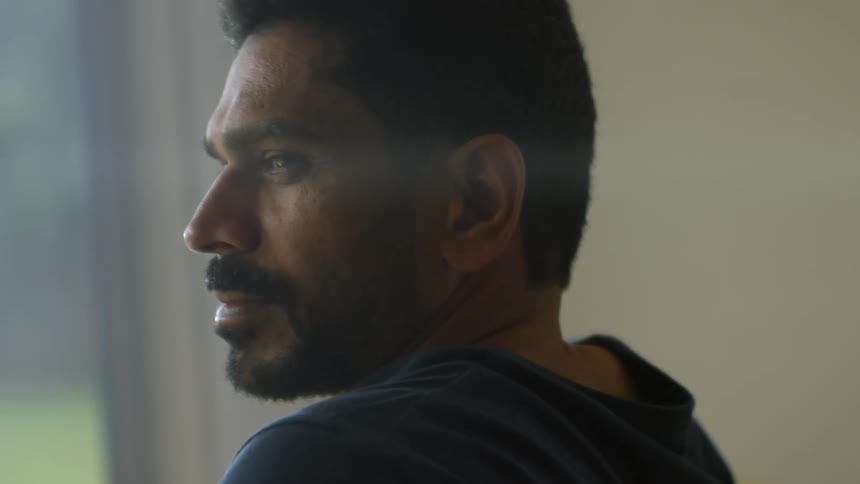 A contemplative man with a beard and mustache looks out a window with a thoughtful expression on his face