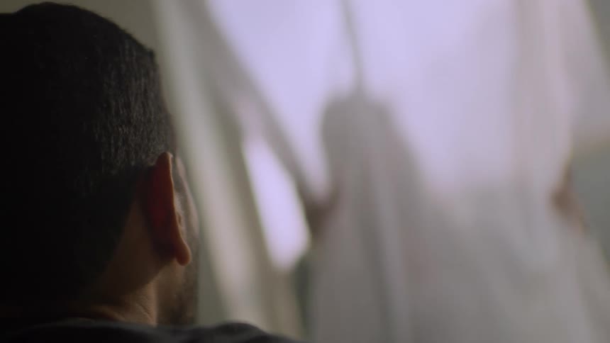 Over-the-shoulder shot of a man watching the mysterious silhouette of a person behind a sheer, white curtain