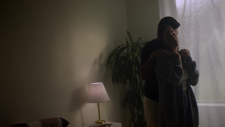 A man tenderly hugs a woman from behind as they stand together in a quiet, dimly lit room by a window