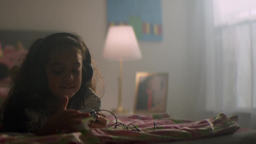 A happy young girl with headphones lies on her bed in a warmly lit room, smiling as she listens to music