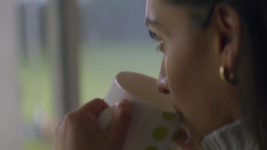 Close-up slow motion shot of a woman pensively drinking from a mug while looking out a window