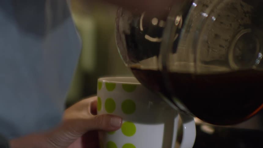 A close-up of fresh, hot coffee being poured from a glass pot into a white and green polka-dotted mug